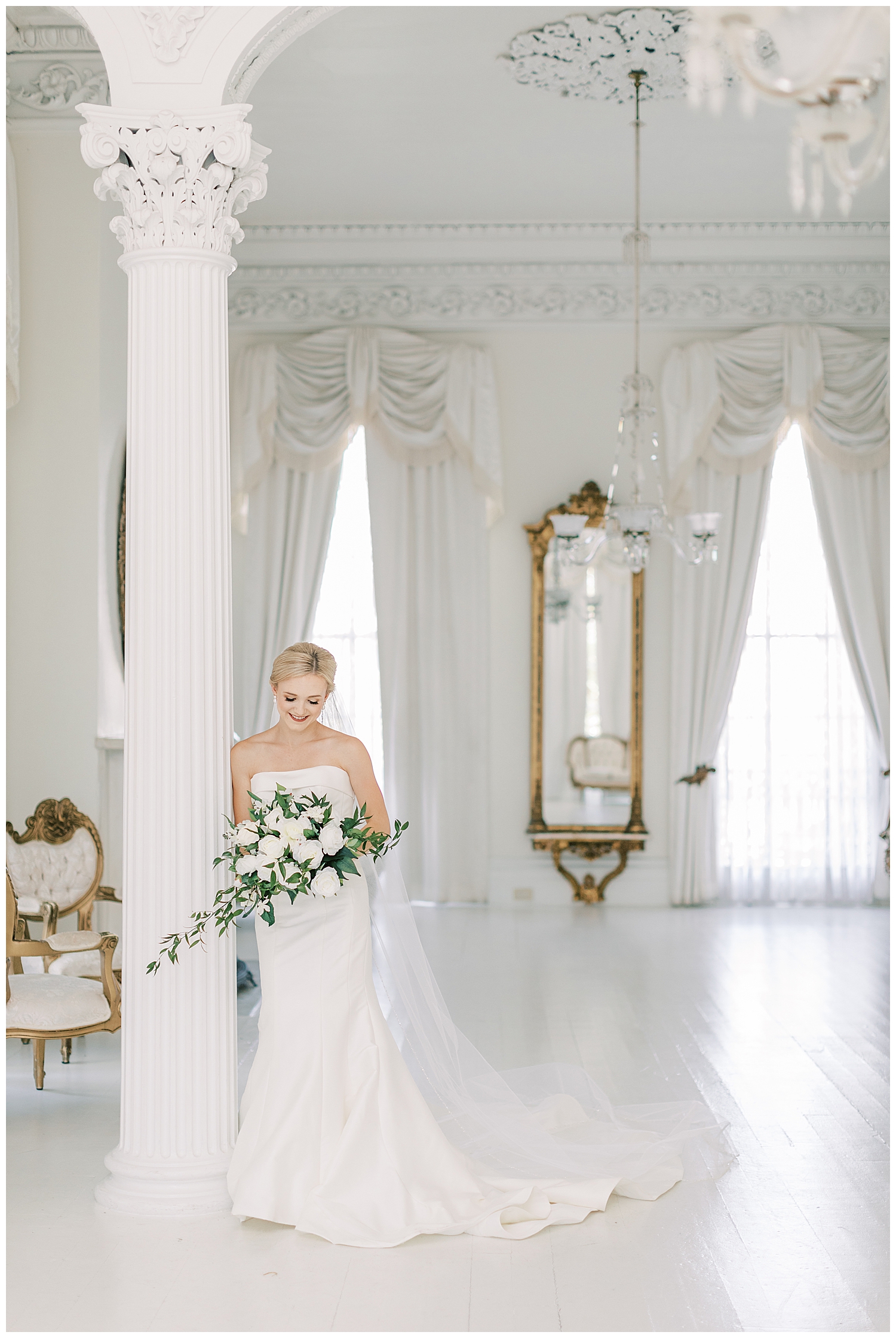A bride holds her bouquet leaning against a large white column in a white room.