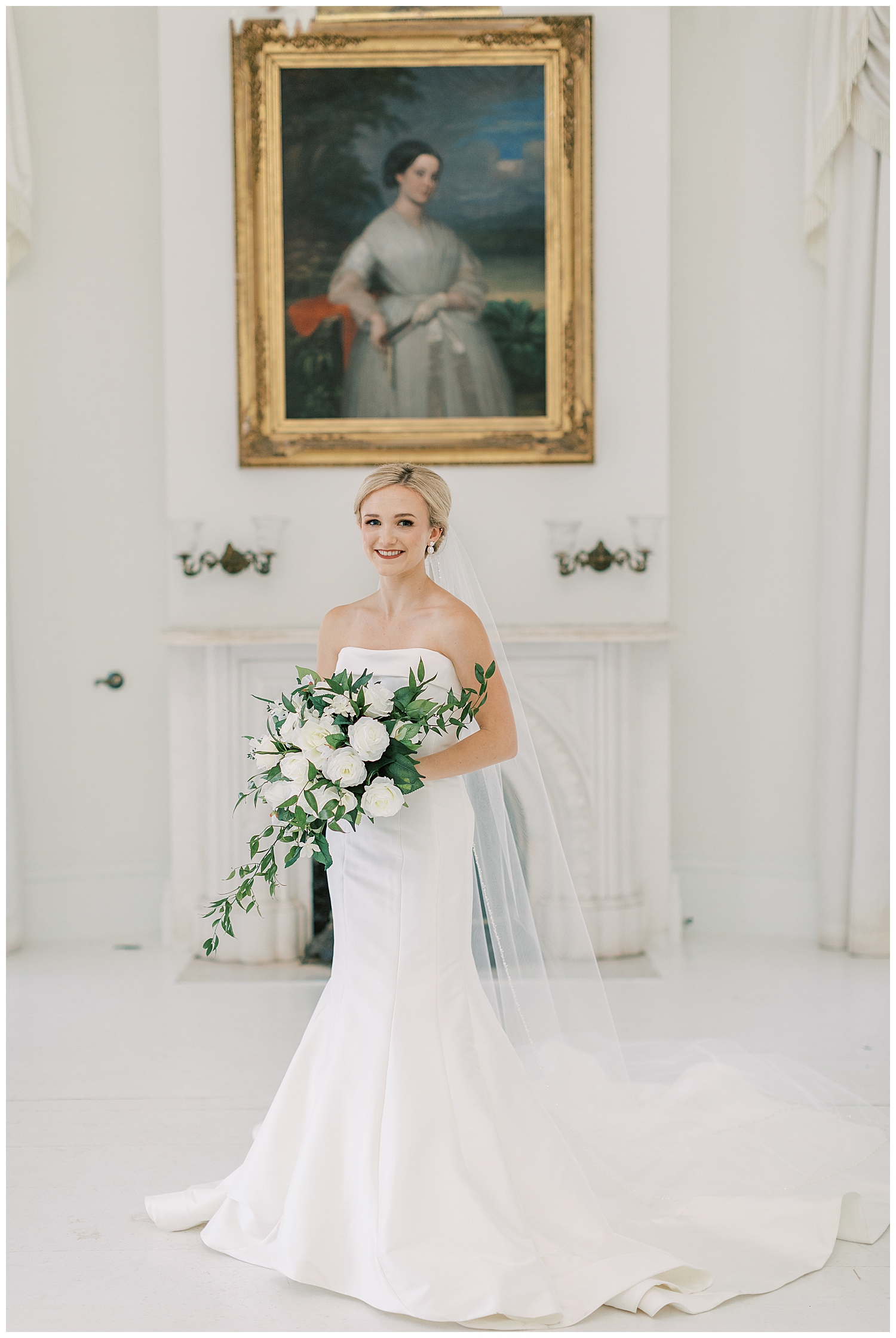 A vintage painting hangs on the wall behind the bride in a white room.