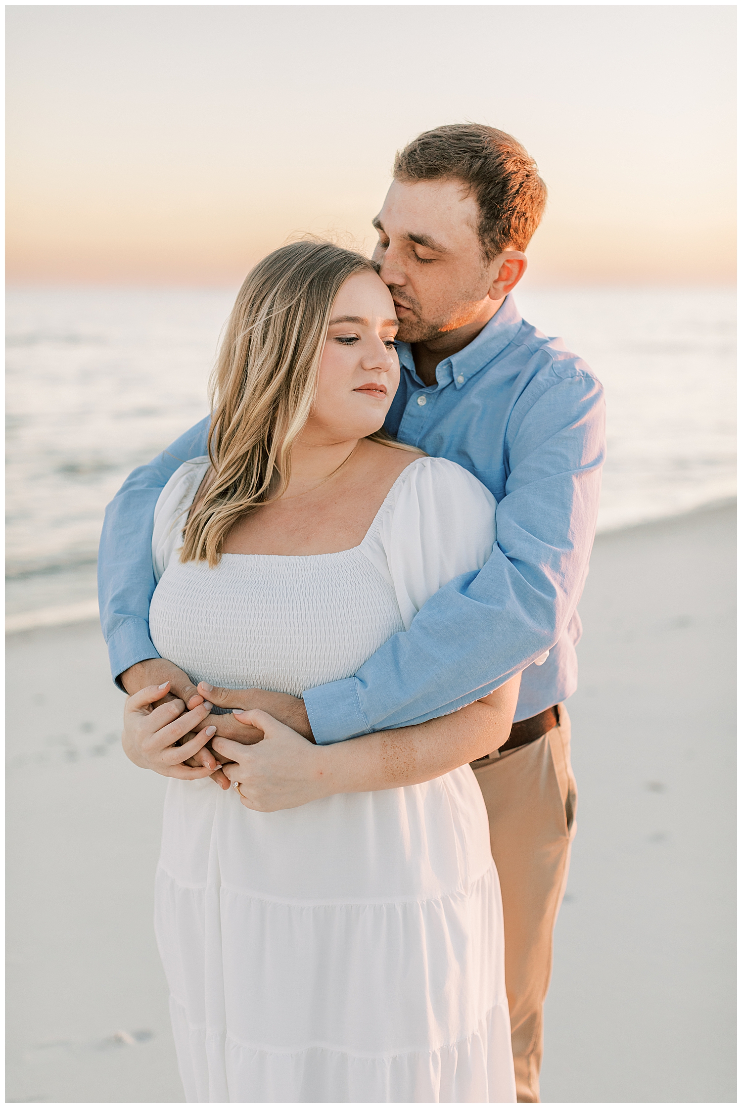 A couple stands on the beach at sunset for their engagement photos.